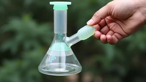 A hand holding a small, clear glass bong with green accents, ideal for a minimalist smoking experience.