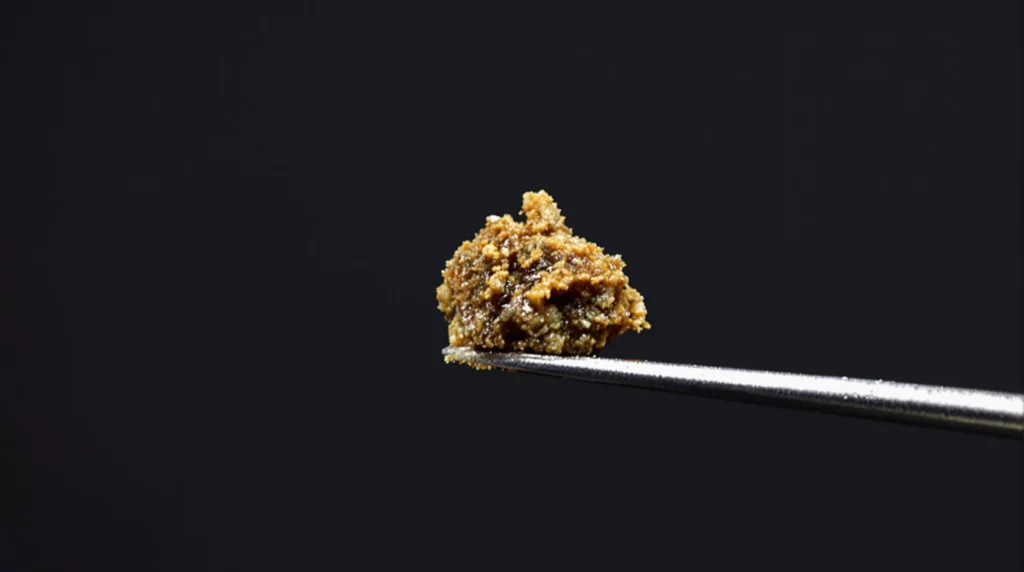 A dab tool holding a small, sticky piece of THCA classic hash, with a dark background to highlight its texture and shine. Soft lighting brings out the natural colors and dense consistency, illustrating the richness and potency of the concentrate.
