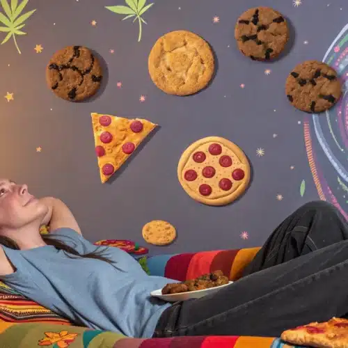 Woman reclining on a couch with floating cookies, pizza slices, and a psychedelic swirl backdrop featuring cannabis leaves.