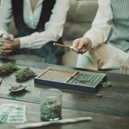 Two people rolling joints on a wooden table with cannabis buds, rolling papers, and grinders in a casual setting